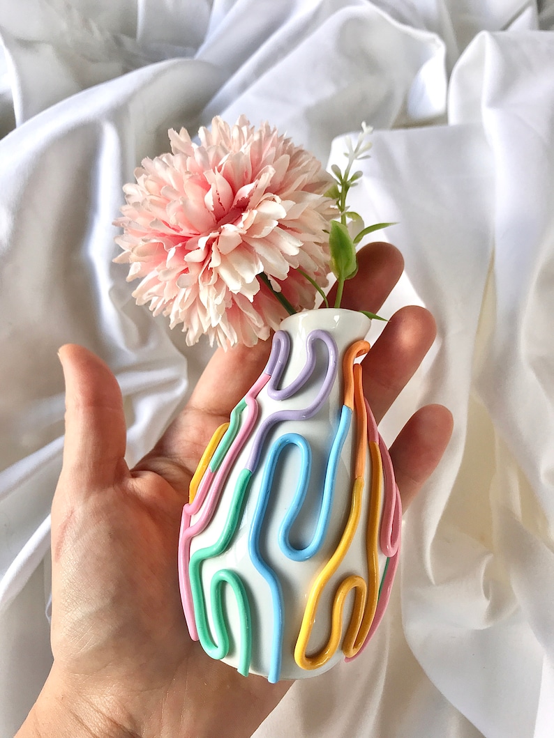 May include: A small white ceramic vase with a colorful abstract design. The vase is decorated with pastel pink, yellow, blue, and green lines.