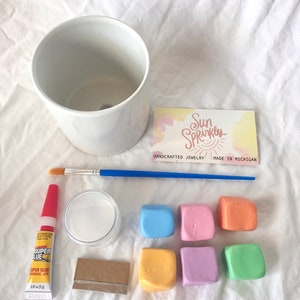 May include: A DIY planter kit with a white ceramic pot, six colorful clay squares, a small bottle of super glue, a small jar of white glue, a paintbrush, and a small piece of wood. The kit also includes a card with the text "Sun Sprinkles, Handcrafted Jewelry, Made in Michigan."