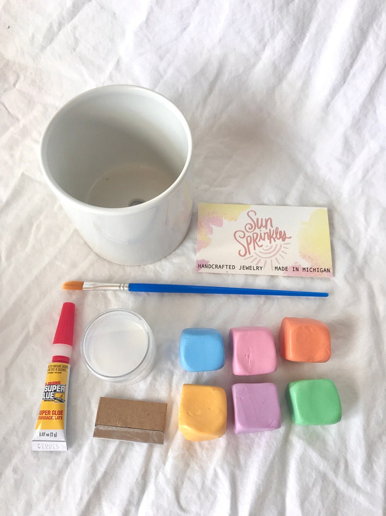 May include: A DIY planter kit with a white ceramic pot, six colorful clay squares, a small bottle of super glue, a small jar of white glue, a paintbrush, and a small piece of wood. The kit also includes a card with the text "Sun Sprinkles, Handcrafted Jewelry, Made in Michigan."