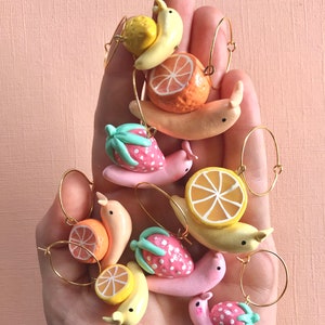 Pu&ograve; includere: Un set di nove ciondoli a forma di lumaca a tema frutta colorati con anelli dorati. Ogni ciondolo presenta un frutto diverso, tra cui una fragola, un'arancia e una banana.