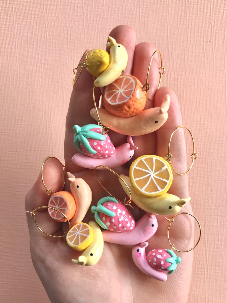 Pu&ograve; includere: Un set di nove ciondoli a forma di lumaca a tema frutta colorati con anelli dorati. Ogni ciondolo presenta un frutto diverso, tra cui una fragola, un'arancia e una banana.