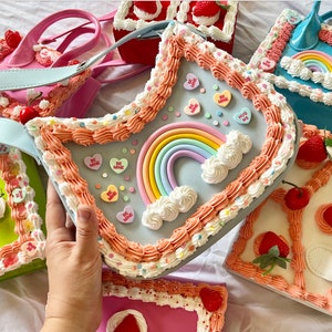 May include: A collection of pastel-colored purses shaped like cakes with frosting, sprinkles, and candy hearts. The purses have rainbow and heart details.