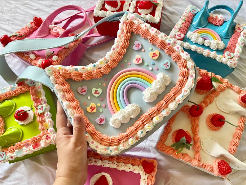 May include: A collection of pastel-colored purses shaped like cakes with frosting, sprinkles, and candy hearts. The purses have rainbow and heart details.
