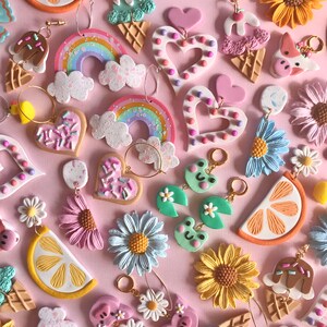 May include: Assortment of colorful polymer clay earrings featuring various designs, including rainbows, hearts, ice cream cones, sunflowers, lemons, and daisies. The earrings are displayed on a pink background.