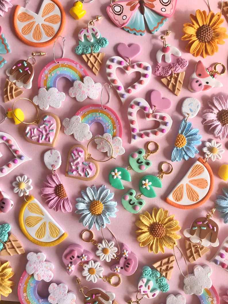 May include: Assortment of colorful polymer clay earrings featuring various designs, including rainbows, hearts, ice cream cones, sunflowers, lemons, and daisies. The earrings are displayed on a pink background.