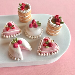 May include: Five miniature clay cakes decorated with pink frosting, white icing, and red and green strawberries. The cakes are arranged on a white oval plate.