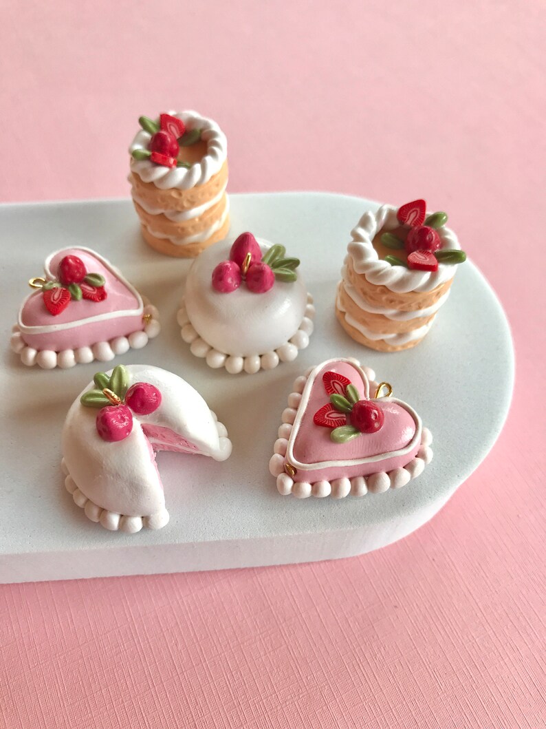 May include: Five miniature clay cakes decorated with pink frosting, white icing, and red and green strawberries. The cakes are arranged on a white oval plate.