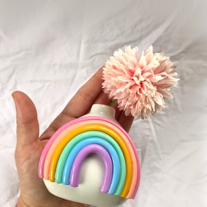 May include: A white ceramic rainbow vase with a pastel rainbow design. The vase is holding a single pink flower.