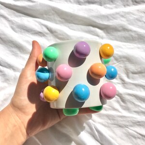 May include: White ceramic mug with pastel colored balls attached to the exterior. The balls are in shades of green, blue, yellow, orange, and purple.