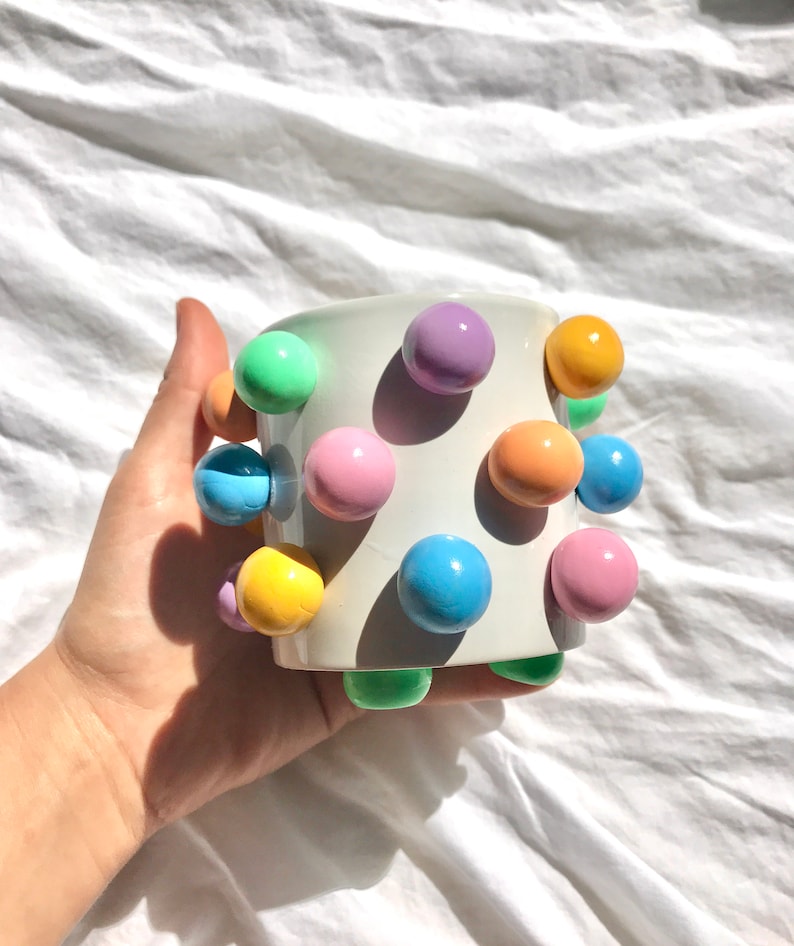 May include: White ceramic mug with pastel colored balls attached to the exterior. The balls are in shades of green, blue, yellow, orange, and purple.