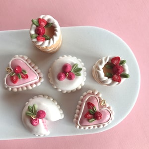 May include: Six miniature cakes decorated with white frosting, red berries, and green leaves. The cakes are arranged on a white plate with a pink background.