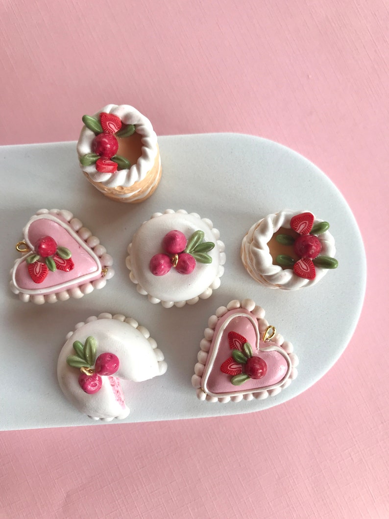 May include: Six miniature cakes decorated with white frosting, red berries, and green leaves. The cakes are arranged on a white plate with a pink background.