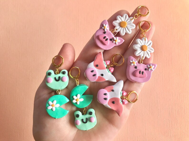 Pu&ograve; includere: Un set di sei orecchini fatti a mano in pasta polimerica con vari design, tra cui una rana, una ninfea, una mucca e un maiale. Gli orecchini sono tutti rosa, verdi e bianchi con accenti dorati.