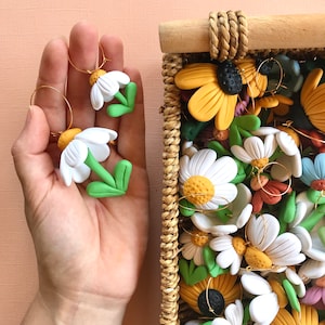 May include: A collection of handmade polymer clay earrings featuring white daisy flowers with green stems and gold hoops. The earrings are displayed in a wicker basket.