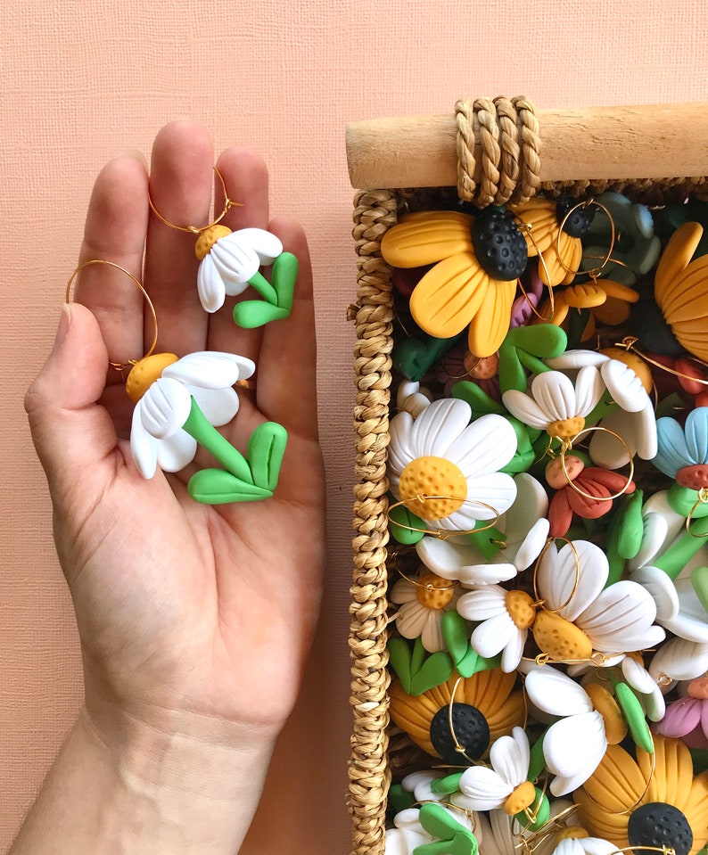 May include: A collection of handmade polymer clay earrings featuring white daisy flowers with green stems and gold hoops. The earrings are displayed in a wicker basket.