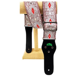 May include: A guitar strap with a decorative paisley pattern in silver, red, and white. The strap has black leather ends and a metal buckle. A green guitar pick is visible in the pick holder.
