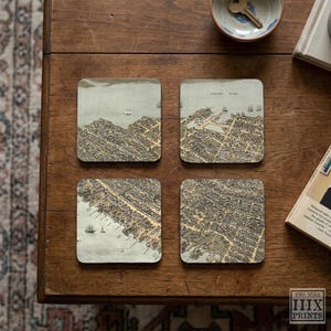 May include: A set of four square coasters featuring detailed vintage-style aerial map illustrations of coastal towns with sailing ships, buildings, and streets in muted sepia, tan, and grey tones, arranged in a two-by-two grid pattern.
