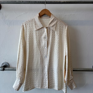 May include: A long-sleeved button-down shirt in a cream color featuring a subtle, repeating checkered jacquard pattern throughout the fabric. The shirt includes a classic collar, a front button placket with matching buttons, and cuffed sleeves.