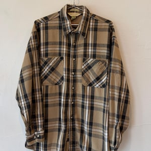 May include: A long-sleeve button-down shirt featuring a plaid pattern in shades of tan, black, and white. The shirt includes a standard collar, two chest pockets with button closures, and a full-length front button placket with matching buttons.
