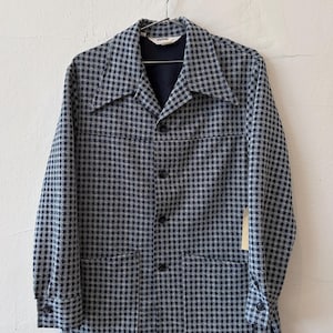 May include: A long-sleeved button-down jacket featuring a black and white houndstooth pattern. The garment includes a pointed collar, four front buttons, two large patch pockets at the waist, and buttoned cuffs. A small tag is visible on the side.