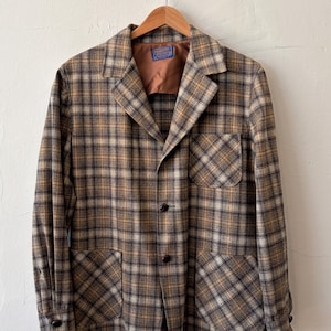 Peut inclure: Un blazer à manches longues avec un motif à carreaux marron, beige et gris. Le vêtement comprend un revers cranté, une poche poitrine, deux poches plaquées inférieures et trois boutons foncés sur le devant. Une étiquette Pendleton est visible.