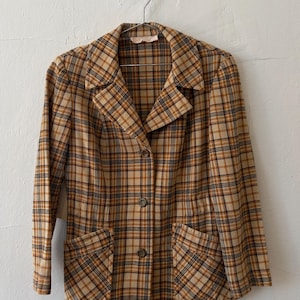 May include: A long-sleeve blazer featuring a tan, brown, and black plaid pattern. The garment includes a notched lapel collar, three front buttons for closure, and two front patch pockets. The blazer is displayed on a metal clothes hanger.