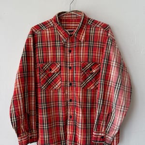 May include: A long-sleeve button-down shirt featuring a red, yellow, and black plaid pattern. The shirt includes a pointed collar, a front button placket, two chest pockets with buttoned flaps, and buttoned cuffs on each long sleeve.