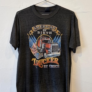 May include: A charcoal grey short-sleeve t-shirt featuring a graphic of a red semi-truck flanked by two American flags. The text reads AMERICAN BY BIRTH and TRUCKER BY CHOICE, with a small emblem that says Trucker Only below the truck.
