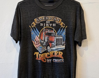 May include: A charcoal grey short-sleeve t-shirt featuring a graphic of a red semi-truck flanked by two American flags. The text reads AMERICAN BY BIRTH and TRUCKER BY CHOICE, with a small emblem that says Trucker Only below the truck.