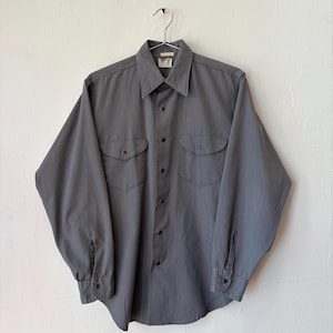 May include: A long-sleeve button-down shirt in a solid slate gray color. The shirt features a pointed collar, a front button placket, two chest pockets with buttoned flaps, and buttoned cuffs. The garment is displayed on a metal hanger.