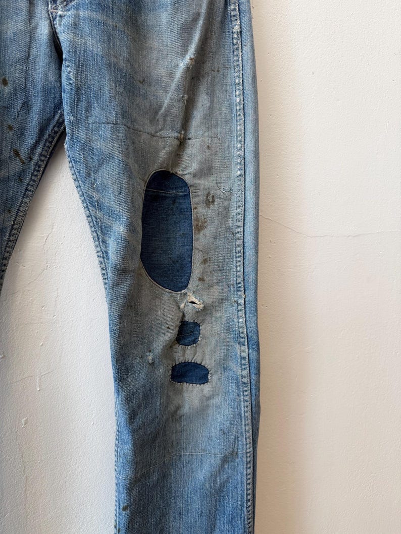 K&ouml;nnte beinhalten: Hellblaue Jeans mit Distressed-Details, einschlie&szlig;lich eines gro&szlig;en Knierisses und Flicken. Die Jeans weisen Gebrauchsspuren auf, mit sichtbarem Ausbleichen und Flecken, was ihnen einen Vintage-Look verleiht. Klassische Jeans im Five-Pocket-Stil.