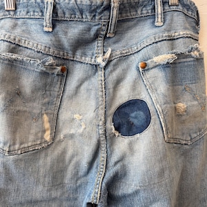 K&ouml;nnte beinhalten: Hellblaue Jeans mit Used-Details, einschlie&szlig;lich zerrissener Taschen und einem runden Flicken auf der R&uuml;ckseite. Die Jeans h&auml;ngen an einem Metallb&uuml;gel und zeigen die Ges&auml;&szlig;taschen und G&uuml;rtelschlaufen.