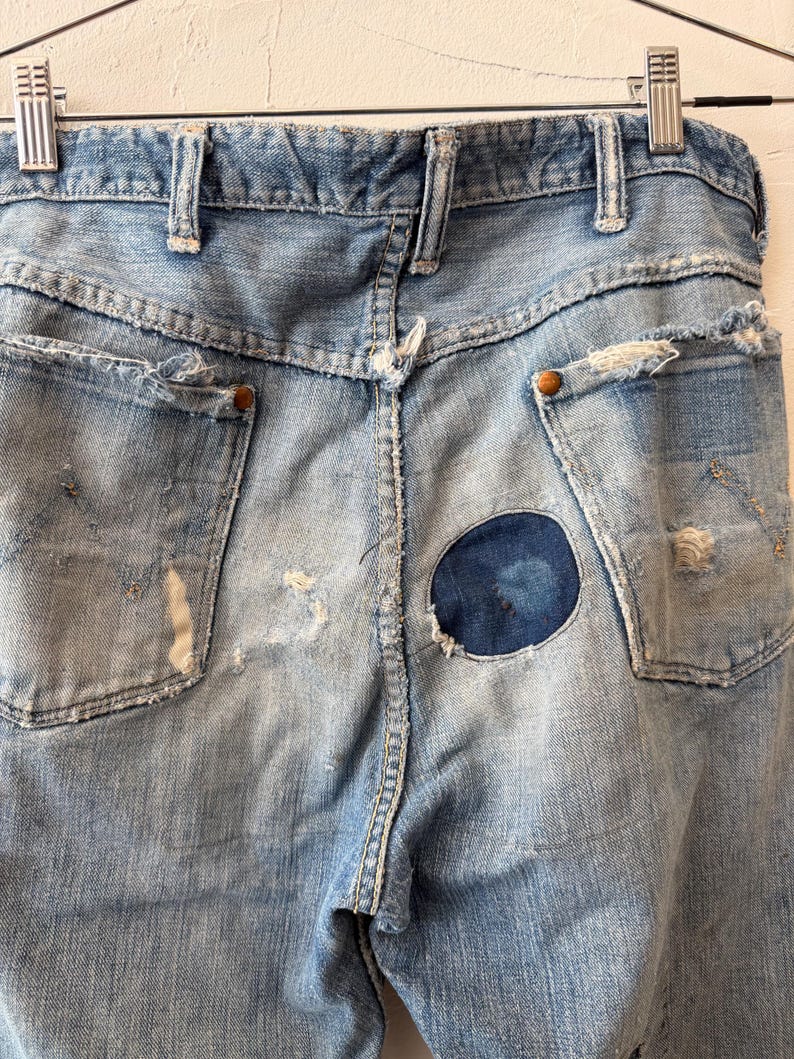 K&ouml;nnte beinhalten: Hellblaue Jeans mit Used-Details, einschlie&szlig;lich zerrissener Taschen und einem runden Flicken auf der R&uuml;ckseite. Die Jeans h&auml;ngen an einem Metallb&uuml;gel und zeigen die Ges&auml;&szlig;taschen und G&uuml;rtelschlaufen.
