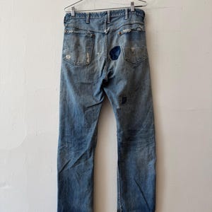 K&ouml;nnte beinhalten: Hellblaue Jeans mit sichtbaren Gebrauchsspuren, Rissen und Flicken. Die Jeans h&auml;ngt an einem Metallb&uuml;gel vor einer wei&szlig;en Wand. Die Ges&auml;&szlig;taschen und der Hosenboden sind zu sehen.