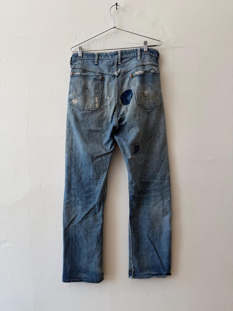 K&ouml;nnte beinhalten: Hellblaue Jeans mit sichtbaren Gebrauchsspuren, Rissen und Flicken. Die Jeans h&auml;ngt an einem Metallb&uuml;gel vor einer wei&szlig;en Wand. Die Ges&auml;&szlig;taschen und der Hosenboden sind zu sehen.