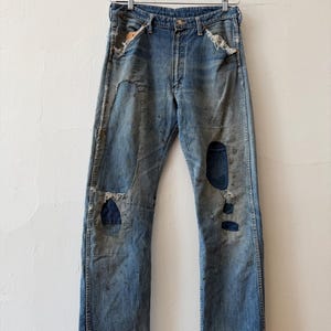 K&ouml;nnte beinhalten: Vintage blaue Jeans aus Denim mit abgenutzten Details, einschlie&szlig;lich Flicken und Rissen. Die Jeans h&auml;ngen an einem Metallb&uuml;gel vor einer wei&szlig;en Wand. Die Jeans haben einen Knopfverschluss und ein klassisches F&uuml;nf-Taschen-Design.
