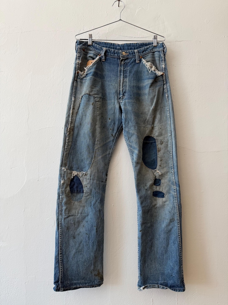 K&ouml;nnte beinhalten: Vintage blaue Jeans aus Denim mit abgenutzten Details, einschlie&szlig;lich Flicken und Rissen. Die Jeans h&auml;ngen an einem Metallb&uuml;gel vor einer wei&szlig;en Wand. Die Jeans haben einen Knopfverschluss und ein klassisches F&uuml;nf-Taschen-Design.