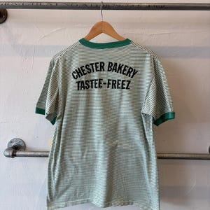 May include: A short-sleeve ringer t-shirt featuring thin horizontal green and white stripes. The shirt has a solid green collar and sleeve cuffs. The back displays black text that reads CHESTER BAKERY TASTEE-FREEZ in a bold font.