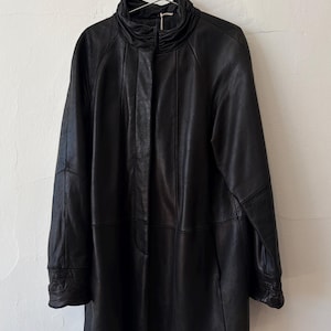 May include: A black leather jacket featuring a high, gathered collar and a concealed front closure. The jacket has long sleeves with cuffed wrists and vertical paneling throughout the body, creating a structured, straight-cut silhouette.