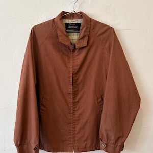 May include: A solid rust-colored zip-up jacket featuring a pointed collar, long sleeves with buttoned cuffs, and two front pockets. The interior is lined with a light-colored plaid fabric, and a black label with the text Sportswear is visible.