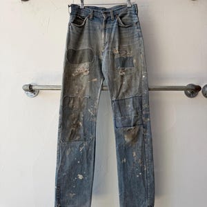 May include: Light blue denim jeans featuring a distressed finish with multiple patches, frayed areas, and scattered paint splatters. The jeans have a classic five-pocket design, a button fly, and belt loops, hanging from a wooden hanger.