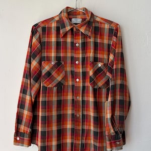 May include: A long-sleeve button-down shirt featuring a plaid pattern in shades of orange, red, black, and tan. The shirt includes a pointed collar, two chest pockets with button closures, and white buttons running down the front placket.