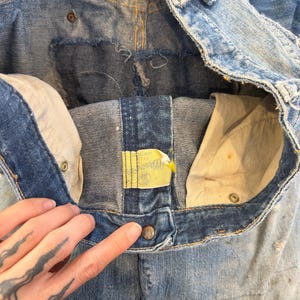 K&ouml;nnte beinhalten: Nahaufnahme einer Vintage-Jeans aus blauem Denim, die das Innere und die Details des Bunds, der Taschen und des Knopfverschlusses zeigt. Die Jeans hat ein abgenutztes, abgenutztes Aussehen mit sichtbaren Gebrauchsspuren.