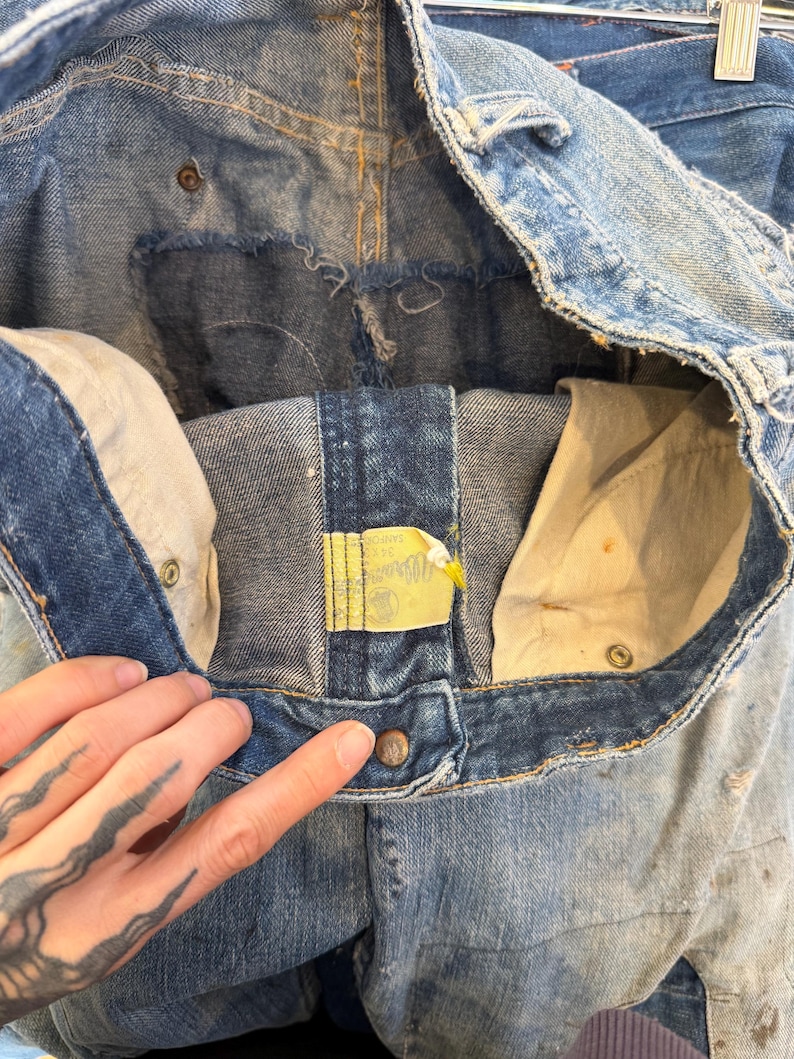 K&ouml;nnte beinhalten: Nahaufnahme einer Vintage-Jeans aus blauem Denim, die das Innere und die Details des Bunds, der Taschen und des Knopfverschlusses zeigt. Die Jeans hat ein abgenutztes, abgenutztes Aussehen mit sichtbaren Gebrauchsspuren.