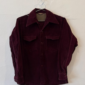 May include: A long-sleeve button-down shirt in deep burgundy corduroy fabric. The shirt features a pointed collar, two chest pockets with buttoned flaps, and matching buttons down the front placket and on the cuffs of the sleeves.