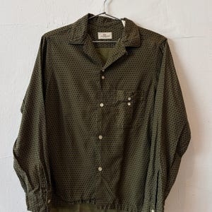 May include: A long-sleeve button-down shirt in olive green with a repeating dark geometric pattern. The shirt features a collared neckline, a single chest pocket with two small buttons, and a label inside the collar that reads Van Heusen.
