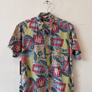 May include: A short-sleeve button-down shirt featuring a vibrant, multi-colored geometric and floral pattern. The design includes red, blue, and lime green shapes, squares, and leaf motifs on a textured fabric with a standard collar and front buttons.