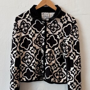 May include: A black and white long-sleeve cardigan featuring a repeating geometric floral pattern. The garment has a solid black velvet-textured collar, a front button closure, and small white bead accents integrated into the embroidered design.