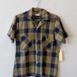 May include: A short-sleeve button-down shirt featuring a plaid pattern in olive green and navy blue with thin brown accent lines. The shirt has a pointed collar, two front chest pockets, and white buttons down the center front placket.