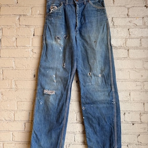 Può includere: Pantaloni da lavoro in denim blu sbiadito con dettagli usurati, tra cui fori e segni di usura. I pantaloni hanno un classico design a cinque tasche e una chiusura con bottoni. Sono appesi a una gruccia di legno contro un muro di mattoni.